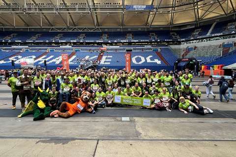 Eine große Gruppe von Personen in grünen T-Shirts steht und sitzt in einer Sportarena, vorne liegen zwei Maskottchen im Drachen- und Bärenkostüm, einige Personen halten ein Banner mit der Aufschrift „B2Run 2025“.