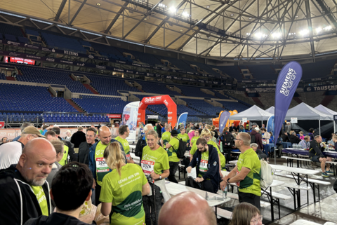 Mehrere Personen in grünen T-Shirts stehen und sitzen an Bierzeltgarnituren in einer großen überdachten Arena mit leeren Tribünen, im Hintergrund sind Zelte, Werbebanner und ein roter Torbogen.