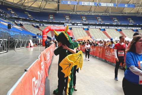 Eine Person in einem grünen Drachenkostüm umarmt eine andere Person auf einer Laufveranstaltung in einem Stadion, während weitere Personen im Hintergrund laufen.