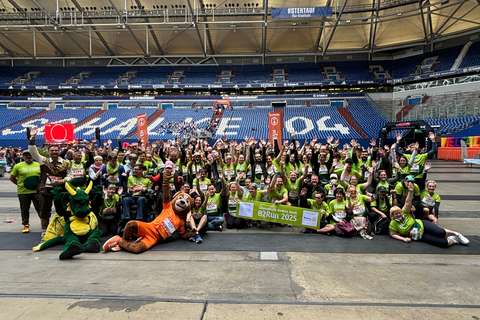 Eine große Gruppe von Personen in grünen T-Shirts steht und sitzt in einer Sportarena, einige halten ein Banner mit der Aufschrift „B2Run 2025“, vorne sind zwei Maskottchen im Drachen- und Tierkostüm.