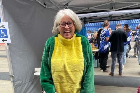 Eine Person mit Brille trägt ein grünes und gelbes Tierkostüm und steht in einer Halle, im Hintergrund mehrere Personen an einem Stand.