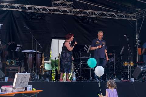 Eine Person und ein Kind mit zwei Luftballons stehen vor einer Bühne, auf der zwei Personen mit Mikrofonen stehen.