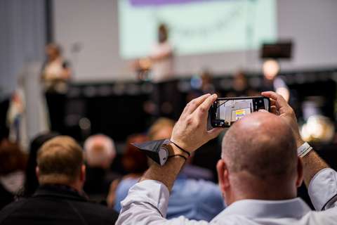 Eine Person hält ein Smartphone hoch und macht ein Foto oder Video von einer Veranstaltung, während mehrere Personen im Publikum sitzen.