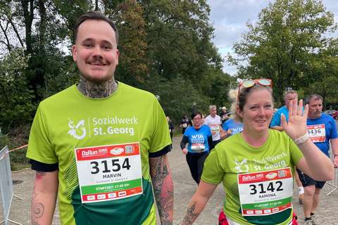 Zwei Personen mit Startnummern und grünen T-Shirts nehmen an einem Lauf teil, im Hintergrund weitere laufende Personen.