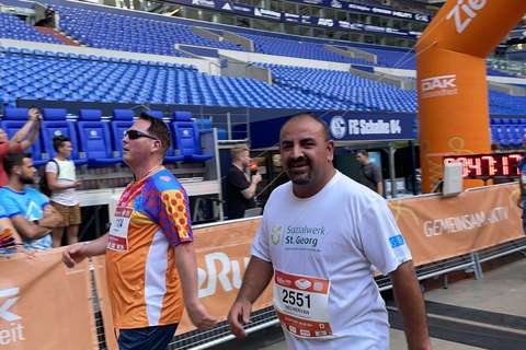 Zwei Personen gehen nebeneinander in einem Stadion an einem orangefarbenen Zielbogen vorbei.