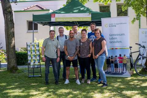 Sieben Personen stehen auf einer Wiese vor einem grünen Pavillon und mehreren Informationsaufstellern.