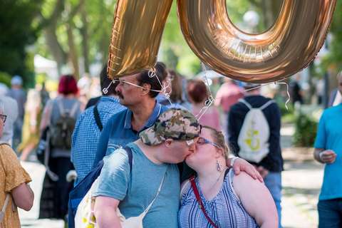 Zwei Personen küssen sich unter einer großen, goldenen Ballonzahl „70“ im Freien, umgeben von weiteren Menschen.
