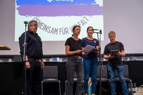 Vier Personen stehen nebeneinander vor Mikrofonen auf einer Bühne, im Hintergrund eine Leinwand mit der Aufschrift „Gemeinsam für Vielfalt“.