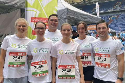 Sechs Personen in Sportkleidung mit Startnummern stehen nebeneinander vor einem grünen Banner mit der Aufschrift „Sozialwerk St. Georg“ in einer großen Halle.