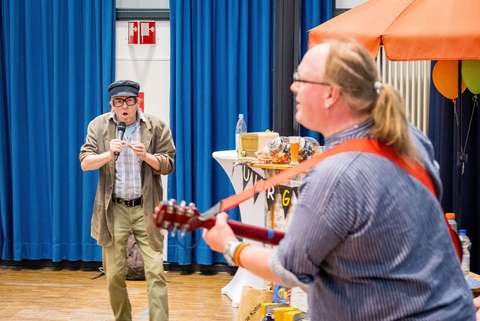 Zwei Personen in einem Raum, eine Person hält ein Mikrofon, die andere spielt Gitarre, im Hintergrund ein Tisch mit verschiedenen Gegenständen und ein orangefarbener Sonnenschirm.