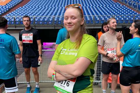 Eine Person mit grünem Laufshirt und Startnummer steht mit verschränkten Armen vor mehreren weiteren Personen in Sportkleidung in einem Stadion.