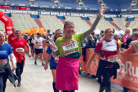 Mehrere Personen mit Startnummern gehen in einem Stadion, eine Person in der Mitte trägt ein grünes Shirt, eine pinke Schürze und hält eine Wasserflasche hoch.