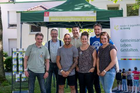 Sieben Personen stehen vor einem grünen Pavillon und einem Werbebanner im Freien.