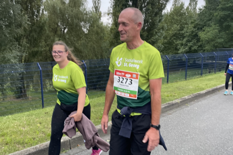 Zwei Personen in Sportkleidung gehen auf einer Straße, beide tragen grüne T-Shirts mit der Aufschrift „Sozialwerk St. Georg“ und Startnummern.