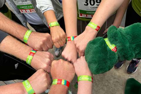Mehrere Personen halten ihre Fäuste zusammen, alle tragen grün-rote Armbänder mit der Aufschrift „B2Run 2025“, eine Person trägt ein grünes Plüschkostüm.