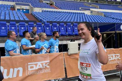 Eine Person läuft in Sportkleidung mit Startnummer durch ein Stadion, im Hintergrund stehen mehrere Personen an einer Absperrung.