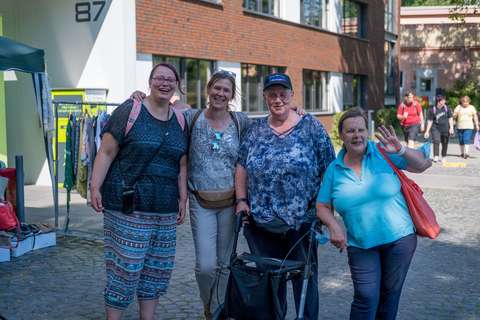 Vier Personen stehen nebeneinander auf einer gepflasterten Straße, eine Person benutzt einen Rollator, im Hintergrund sind weitere Personen und ein Gebäude zu sehen.