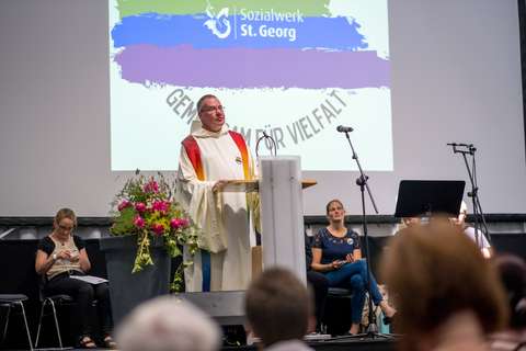 Eine Person im liturgischen Gewand steht an einem Rednerpult, daneben ein großer Blumenstrauß, im Hintergrund sitzen mehrere Personen vor einer Leinwand mit dem Logo „Sozialwerk St. Georg“.
