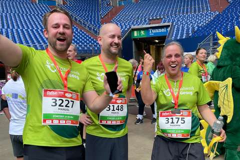 Drei Personen mit grünen Laufshirts und Startnummern stehen lachend nebeneinander in einem Stadion.
