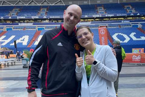 Zwei Personen stehen in einem Stadion, eine Person trägt eine schwarze Jacke mit Manchester-United-Logo, die andere Person zeigt beide Daumen nach oben.