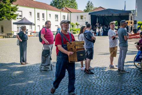 Eine Person mit rotem Shirt und blauer Latzhose trägt eine Drehorgel, mehrere weitere Personen stehen auf einem gepflasterten Platz, im Hintergrund eine Bühne.