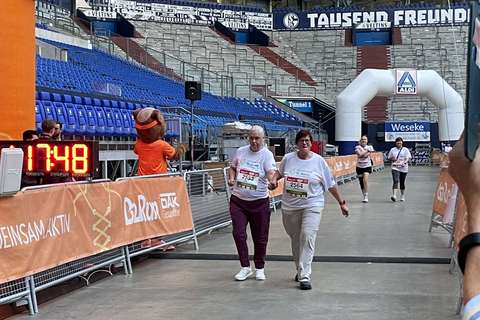 Zwei Personen überqueren gemeinsam eine Ziellinie unter einem orangefarbenen Bogen in einem Stadion.