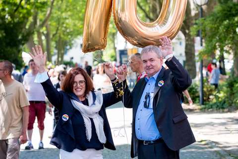 Zwei Personen halten goldene Luftballons in Form der Zahl 70 und heben eine Hand.