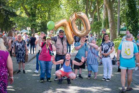 Eine Gruppe von Personen steht und hockt auf einem Weg im Park, einige halten große goldene Luftballons in Form der Zahl 70 und einen grünen Ballon.