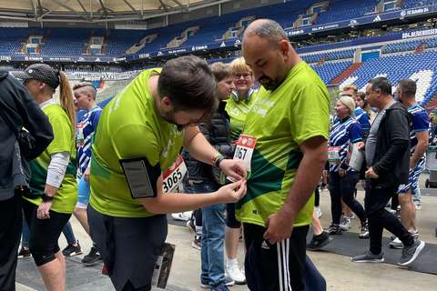 Eine Person befestigt einer anderen Person eine Startnummer an einem grünen T-Shirt, umgeben von weiteren Personen in Sportkleidung in einem Stadion.