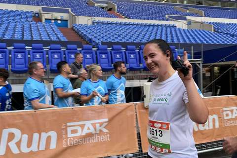 Eine Person mit Startnummer läuft in einem Stadion und hält einen Gegenstand in der Hand, im Hintergrund stehen mehrere Personen vor leeren blauen Sitzreihen.