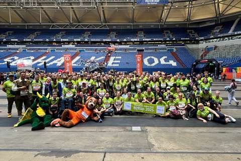 Eine große Gruppe von Personen in grünen T-Shirts steht und sitzt in einem Stadion, vorne liegen zwei Maskottchen im Drachen- und Hundekostüm, einige Personen halten ein Banner mit der Aufschrift „B2Run 2025“.