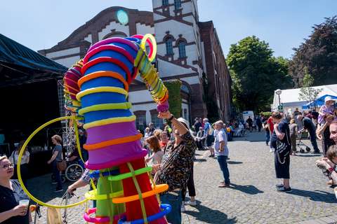 Mehrere Personen stehen auf einem gepflasterten Platz vor einem historischen Gebäude, eine Person trägt ein auffälliges, buntes Kostüm mit Ringen und Schläuchen, während andere zuschauen.