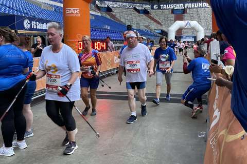 Mehrere Personen überqueren mit Walkingstöcken eine Ziellinie unter einem orangefarbenen Torbogen in einem Stadion.