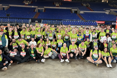 Eine große Gruppe von Personen in grünen T-Shirts mit Startnummern sitzt und steht in einer Halle vor leeren Tribünen.