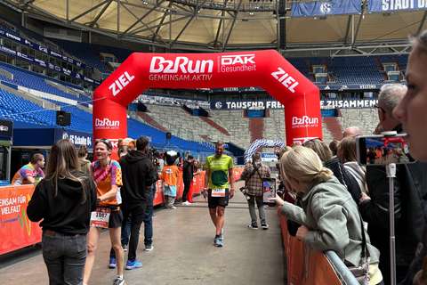 Mehrere Personen befinden sich in einem Stadion an einer Ziellinie unter einem roten Bogen mit der Aufschrift „B2Run“, während Zuschauende am Rand stehen und fotografieren.