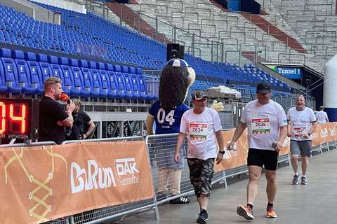 Drei Personen mit Startnummern gehen in einem Stadion, daneben steht eine Person im Maskottchenkostüm mit der Nummer 04 auf dem Rücken.