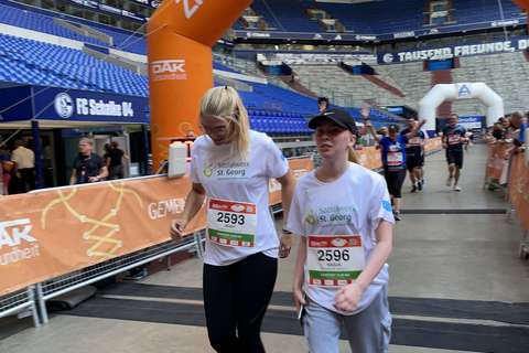 Zwei Personen mit Startnummern und weißen T-Shirts gehen unter einem orangefarbenen Zielbogen in einem Stadion.