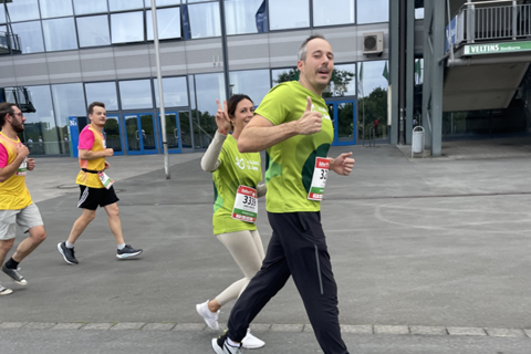 Mehrere Personen laufen in Sportkleidung mit Startnummern auf einer Straße vor einem modernen Gebäude.