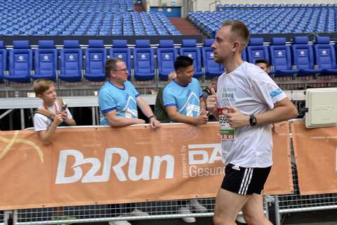 Eine Person läuft in Sportkleidung an einer Absperrung mit B2Run-Banner vorbei, dahinter sitzen und stehen mehrere Personen auf Stadionplätzen.