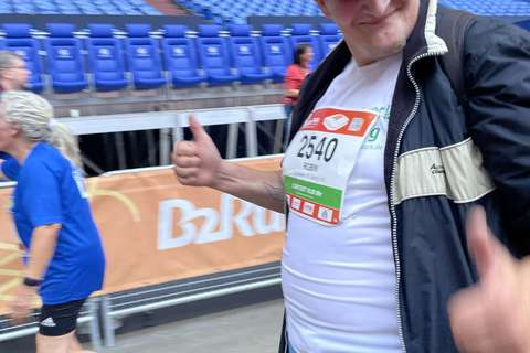Eine Person mit Startnummer und Kappe zeigt beide Daumen hoch, im Hintergrund laufen weitere Personen in einem Stadion.