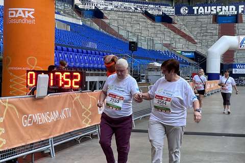 Zwei Personen mit Startnummern überqueren gemeinsam die Ziellinie unter einem orangefarbenen Bogen in einem Stadion.