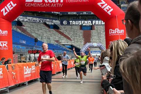 Mehrere Personen laufen durch einen roten Zielbogen bei einem Laufwettbewerb in einem Stadion, während Zuschauer am Rand stehen und fotografieren.
