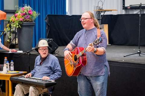 Zwei Personen musizieren, eine spielt Keyboard und trägt einen Bauhelm, die andere steht daneben und spielt Gitarre.