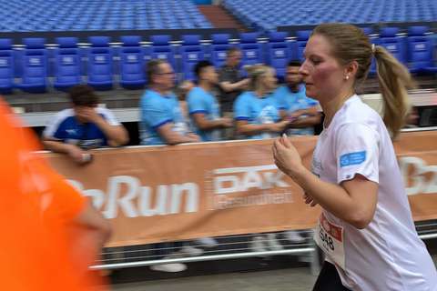 Eine Person läuft in Sportkleidung an einer Absperrung vorbei, im Hintergrund sitzen und stehen mehrere Personen auf blauen Tribünenplätzen.