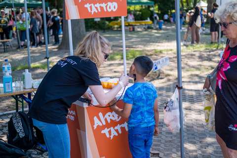 Eine Person trägt einem Kind an einem Stand mit der Aufschrift „Airbrush Tattoo“ ein temporäres Tattoo auf den Hals auf, während eine weitere Person daneben steht.
