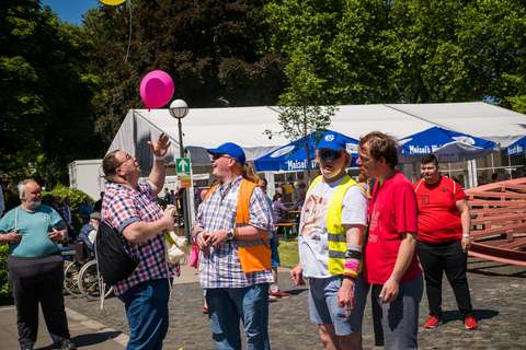 Mehrere Personen stehen im Freien vor einem weißen Zelt, eine Person hält einen pinken Luftballon, andere tragen bunte Westen und T-Shirts.