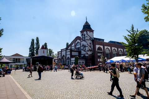 Mehrere Personen gehen und stehen auf einem gepflasterten Platz vor einem großen Gebäude mit Turm, im Hintergrund sind Zelte und eine Bühne aufgebaut.