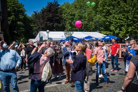 Mehrere Personen stehen im Freien und schauen nach oben, während bunte Luftballons in den Himmel steigen.
