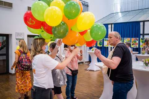 Mehrere Personen stehen in einem Raum mit bunten Luftballons in den Farben Gelb, Grün und Rot.