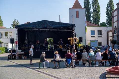Mehrere Personen sitzen und stehen vor einer Bühne im Freien, auf der eine Musikgruppe auftritt; an der Bühne hängt ein goldener Luftballon mit der Zahl 70.